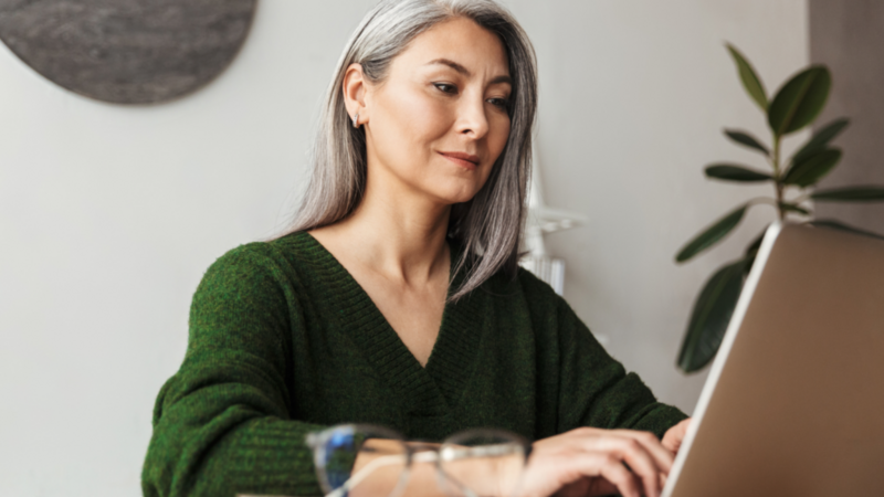 A woman with long gray hair in a green sweater is working on a laptop at a desk. Glasses rest in the foreground.