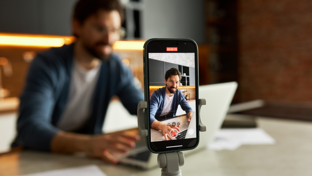 A man sits at a kitchen table with a laptop, smiling while recording a video on a smartphone mounted on a tripod.
