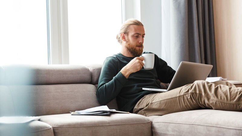A man sitting on a couch, sipping from a mug while using a laptop, learning what a fulfillment center is.