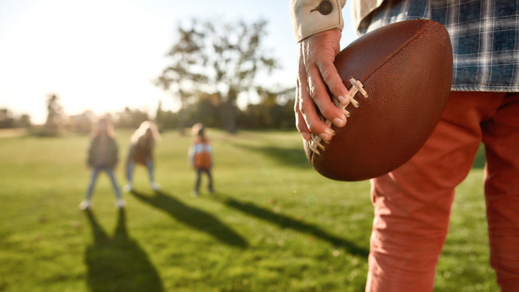 A person in red pants holds a football in the foreground on a sunny day. In the background, three kids stand on a grassy field.