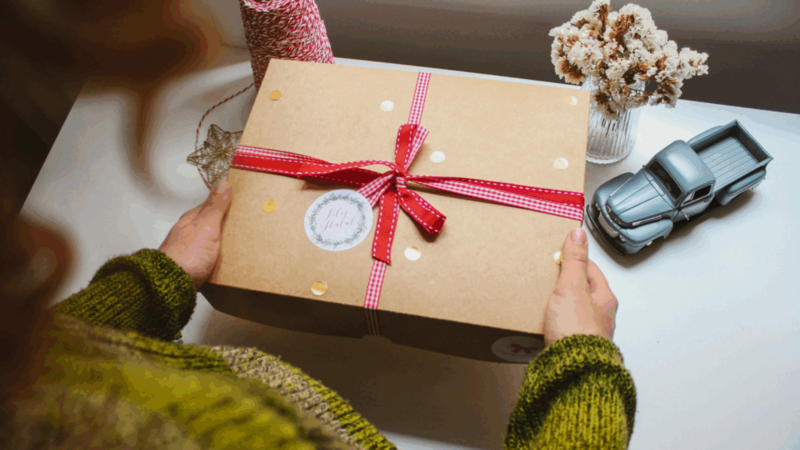 A woman holding a wrapped gift box, smiling, with a table in the background.