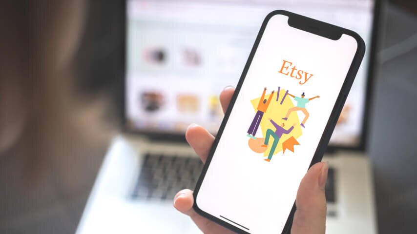 A smartphone displaying an Etsy logo with a blurred laptop in the back; the laptop shows an Etsy page.   