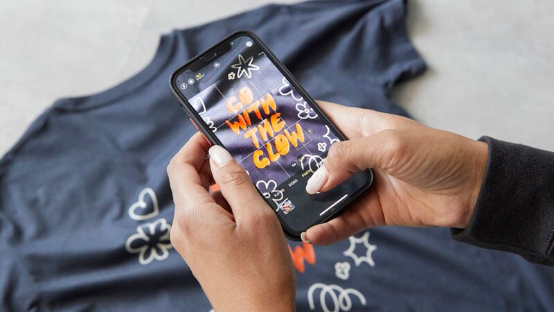 A person taking a picture of a blue “Go with the glow” t-shirt using a smartphone.