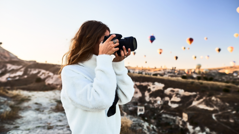 A woman in a white sweater takes a photo with a camera, set against a scenic backdrop of colorful hot air balloons floating in a clear sky at sunrise.