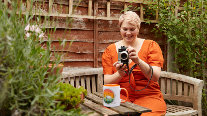 A woman in an orange dress captures a moment with her camera, smiling as she takes a photo outdoors.