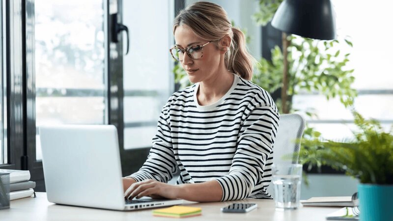 A focused woman in glasses and a striped shirt works on a laptop at a modern desk with plants and a window view.