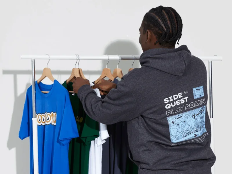 A man in a dark hoodie with retro gaming graphics arranges colorful t-shirts on hangers.