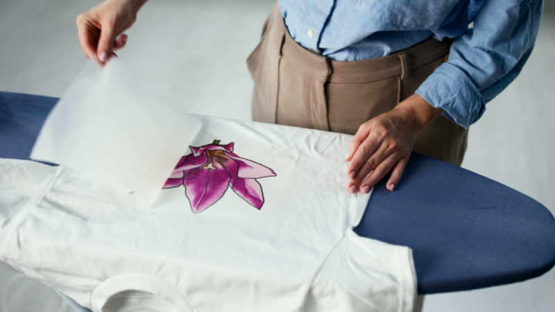 A woman is placing a transfer sheet onto a white t-shirt with a pink flower design on an ironing board. She wears a blue shirt and beige pants.