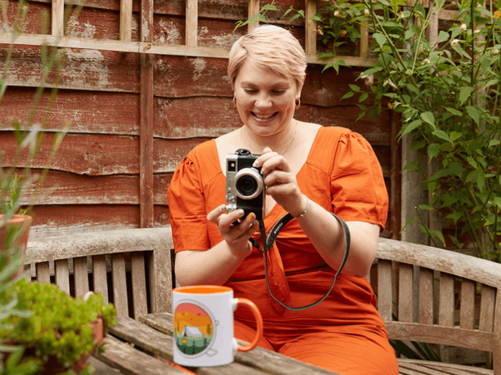 A young woman in an orange dress taking a picture of a custom mug to earn passive income as an artist.