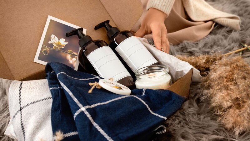 A person's hand reaching into a box filled with skincare products, a candle, and a decorative item.