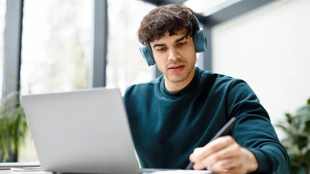 A man wearing headphones is focused on his laptop, engaged in work or a virtual meeting.