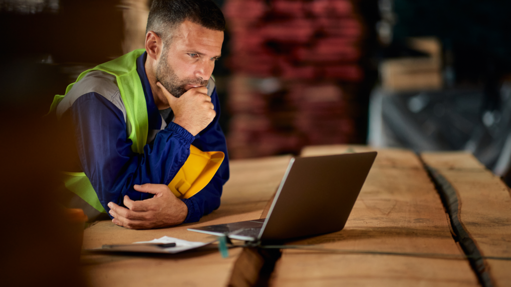 A man sitting at a wooden desk and looking at a laptop screen, thinking.