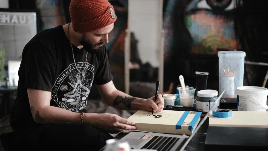 A man wearing a beanie works intently on his laptop at a desk, focused on his task.