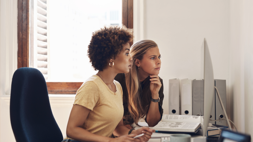 Two women in an office look at a monitor, carefully reading something.