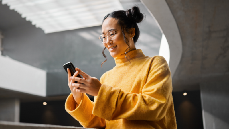A young woman in glasses, wearing a yellow sweater, is looking at her smartphone, searching for TikTok SEO advice.