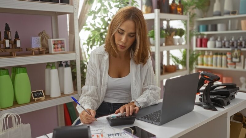 A woman counting something using a calculator, sitting at a desk with a laptop on top of it.