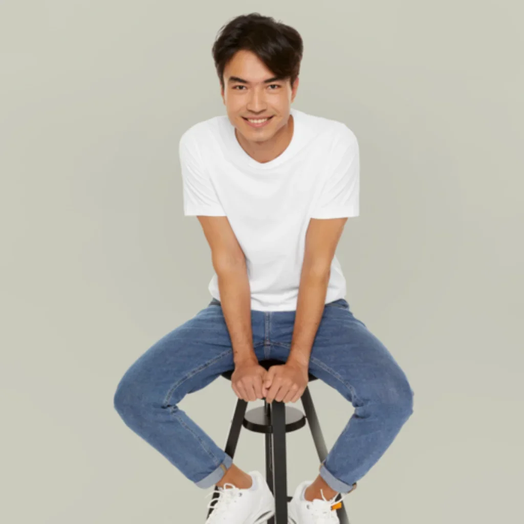 A man in a white t-shirt and jeans sits playfully on a stool against a gray background, smiling warmly at the camera, conveying a relaxed and cheerful mood.