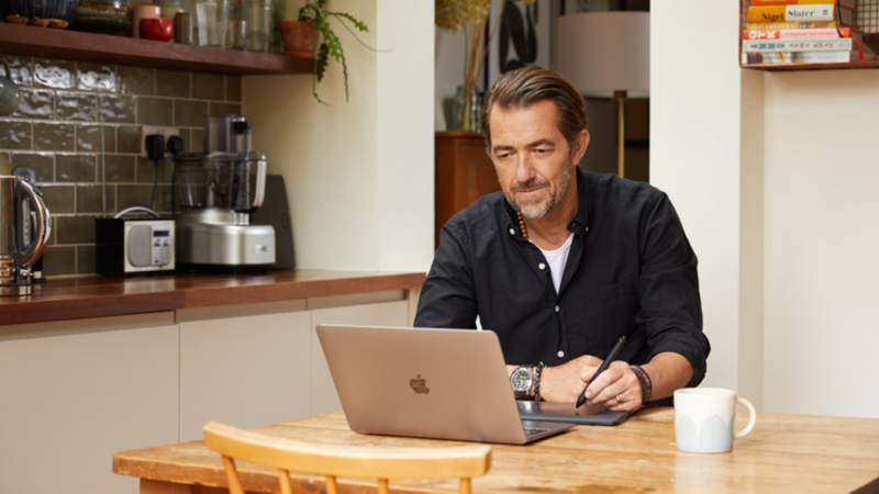 A man in a blue shirt sitting at a kitchen table and using a laptop.
