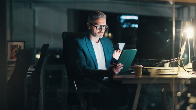A man in a suit sitting at a desk, working on a tablet, searching for Stan Store alternatives in a dimly lit office.