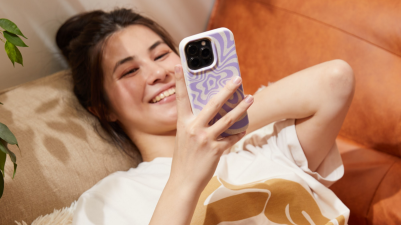 A young woman lies on a couch, smiling as she looks at her phone. She has long hair and is wearing a casual t-shirt.