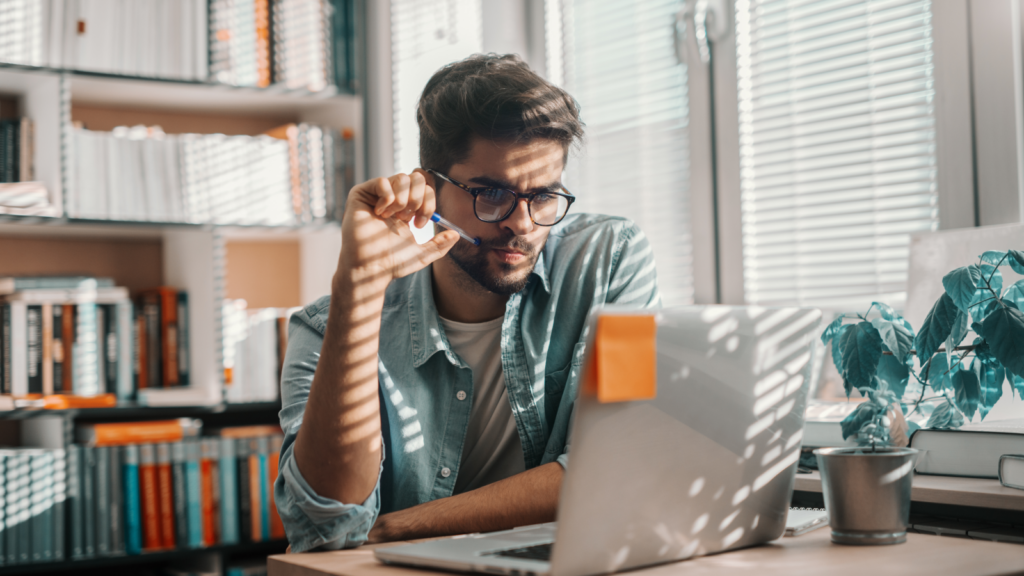 A man in glasses sitting behind a laptop and thinking while looking at the screen.