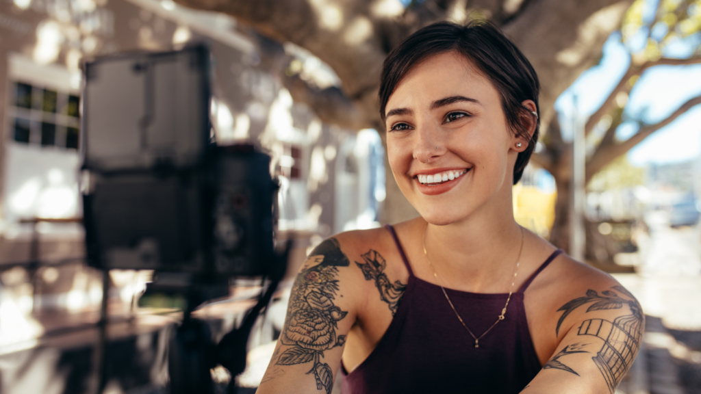 A smiling woman with tattoos filming herself for YouTube.
