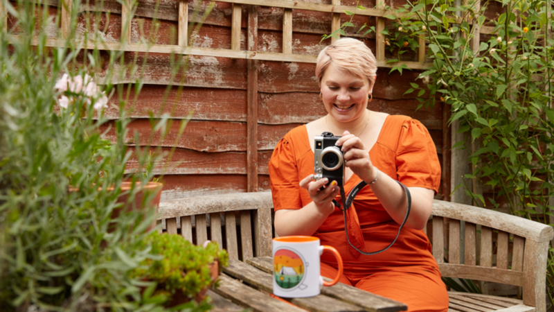 A woman seated on a bench, capturing a customized mug with her camera in a serene outdoor setting.