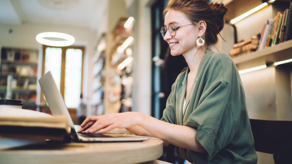 A smiling woman in glasses sitting in a coffee shop and doing freelance work from her laptop.