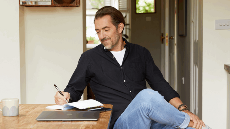 A man in a dark blue t-shirt sitting at a kitchen table, making notes.