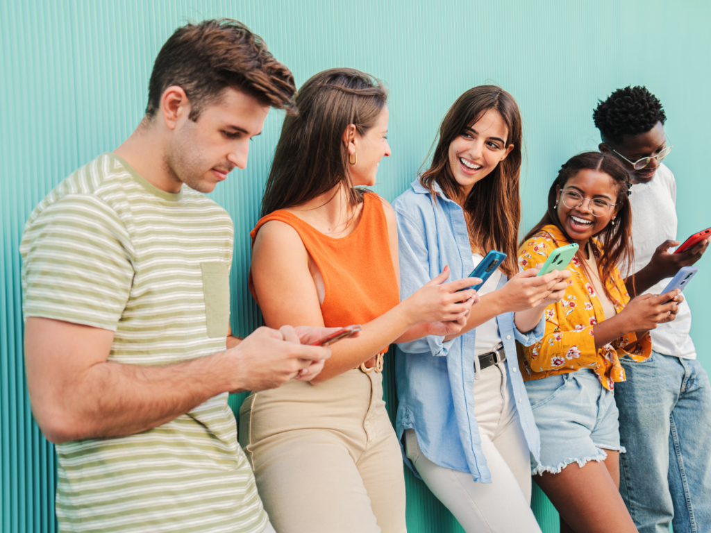 A diverse group of young people standing by a wall, engaged with their phones, enjoying each other's company in a casual setting.
