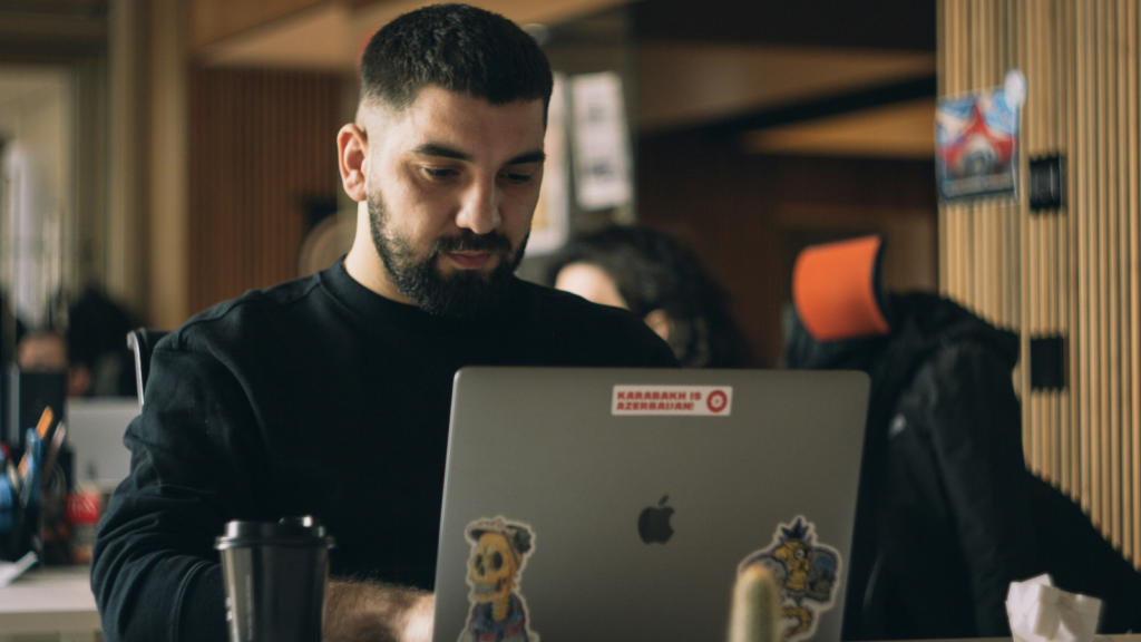 A bearded man in a black hoodie sitting in a cafe and using a laptop.