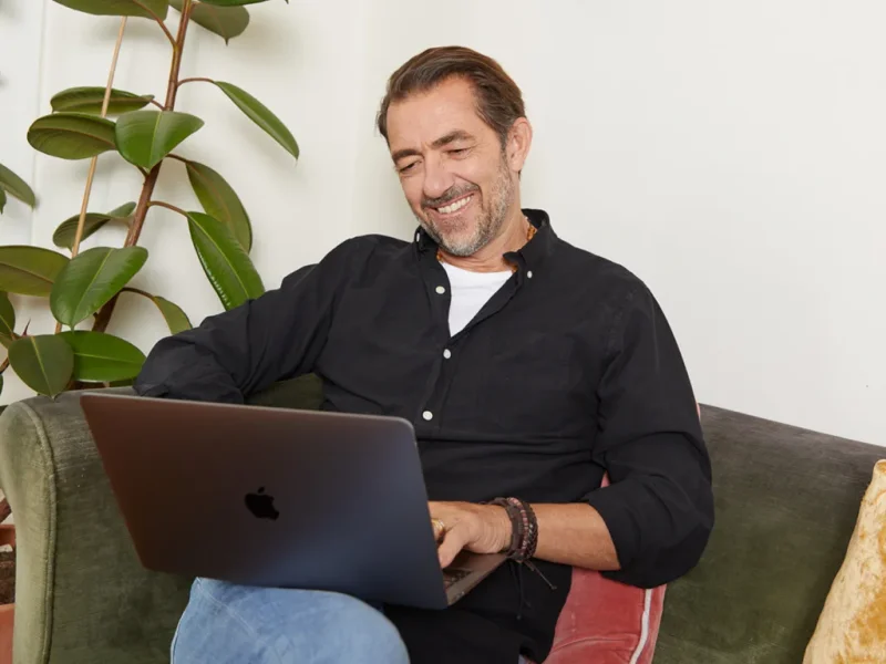 A man sits on a couch, working on his laptop, brainstorming ideas for starting a sticker business.