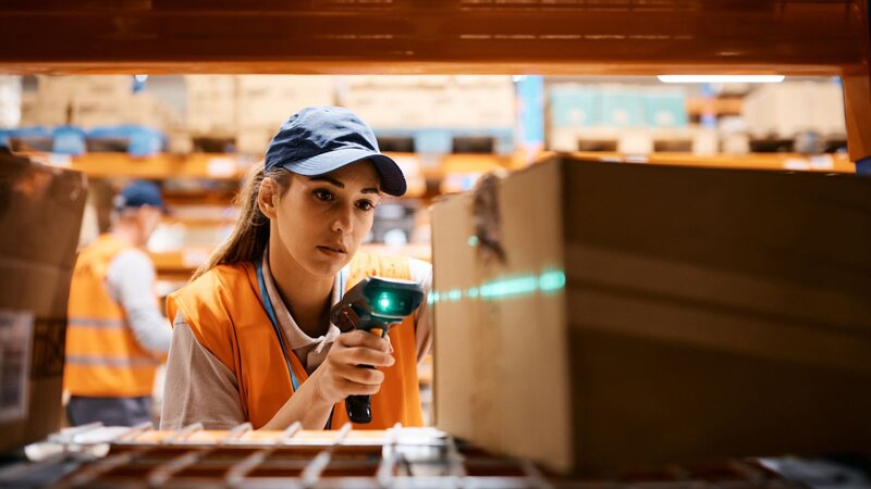 A woman warehouse worker in a cap and orange vest is scanning a box.