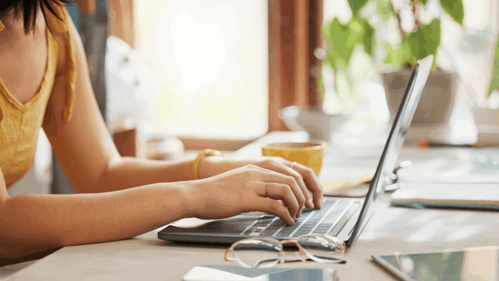 A woman in a yellow top types on a laptop, searching for logo digitizing for embroidery, at a sunlit table.