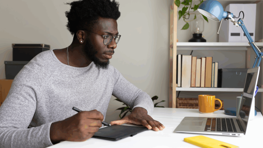 A man wearing glasses is seated at a desk, working on a laptop.