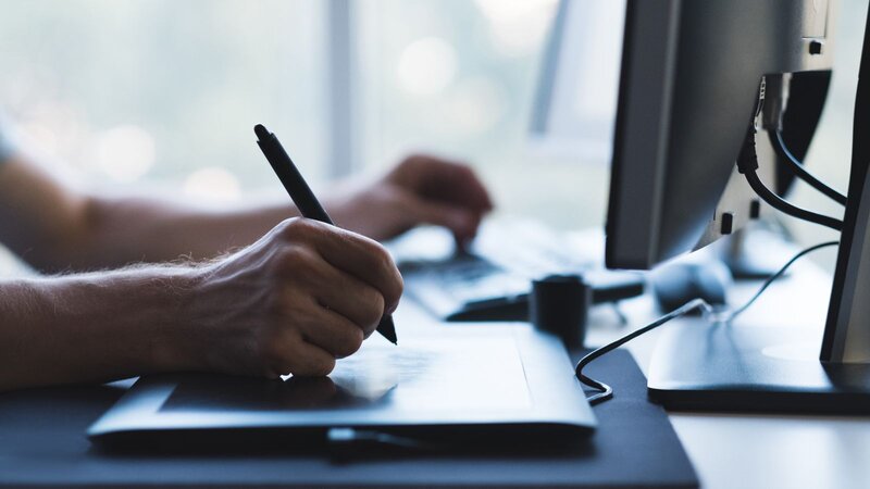 Close-up of a hand drawing on a graphics tablet next to a desktop computer monitor.