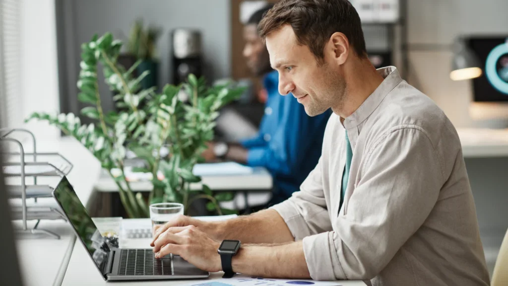 A man in a light shirt sitting at a desk and working from a laptop.