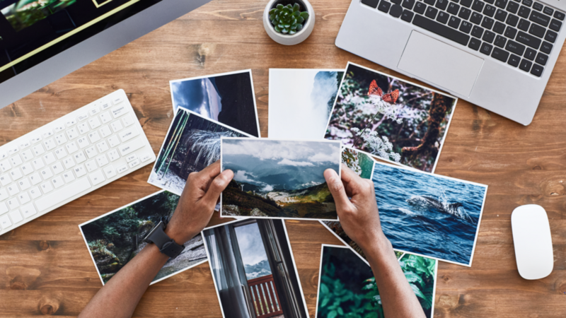 A person’s hands hold a printed photo above a pile of similar photos on a wooden desk. Laptop and PC keyboard nearby.