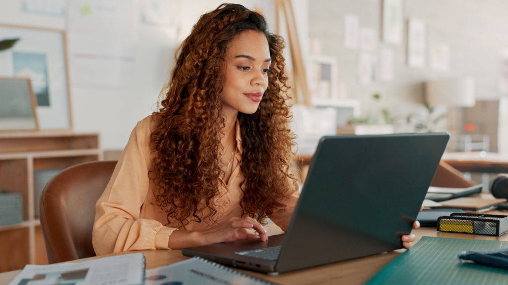 A woman with curly hair, smiling, works intently on a laptop at a desk. The room is decorated with plants and artwork.