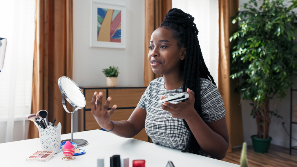 A woman with braided hair in a plaid dress sits at a table with makeup items and a mirror, recording herself using a phone.