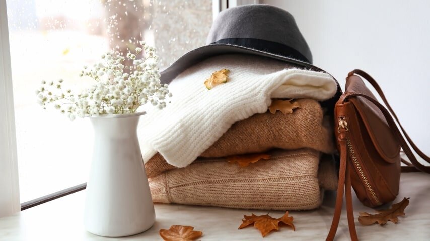 An aesthetic arrangement of autumn sweaters, a wide-brimmed hat, and a messenger bag.