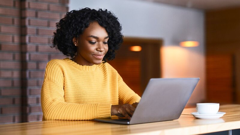 A woman in a yellow sweater uses a laptop to learn how to make money with AI at a wooden table with a cup of coffee nearby.
