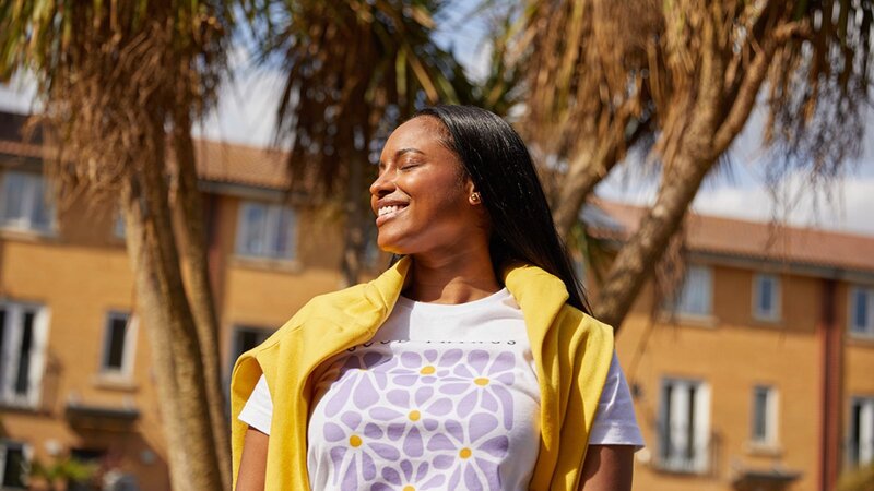 A woman smiling outdoors, wearing a white t-shirt with a floral design and a yellow sweater draped over her shoulders.