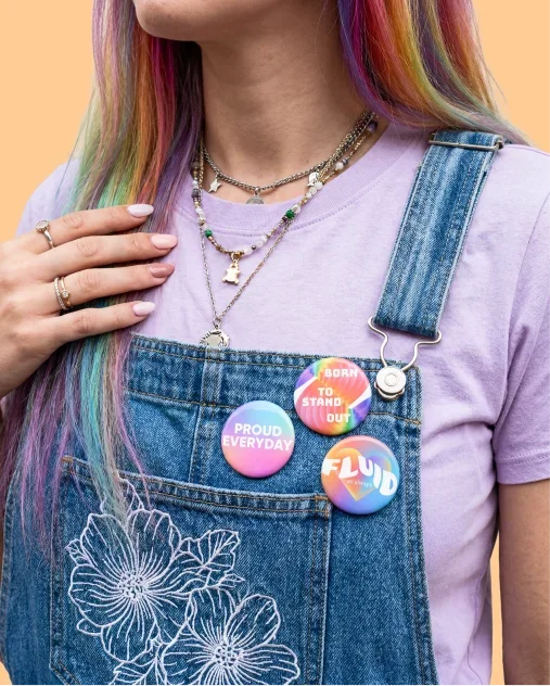 A woman with vibrant rainbow hair wearing buttoned overalls, smiling and standing against a neutral background.