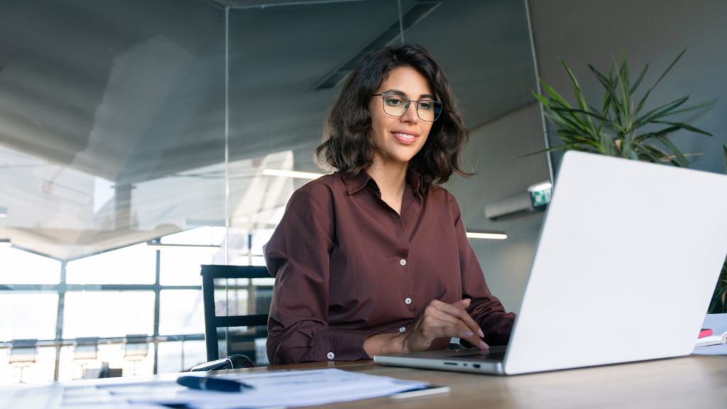 A woman wearing glasses is focused on her laptop, engaged in work at a desk.