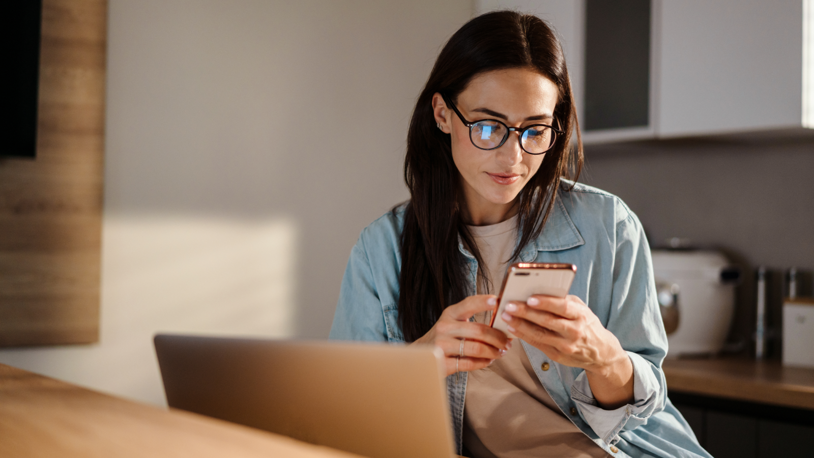 A woman looking at her mobile phone while sitting at a desk in front of a laptop.