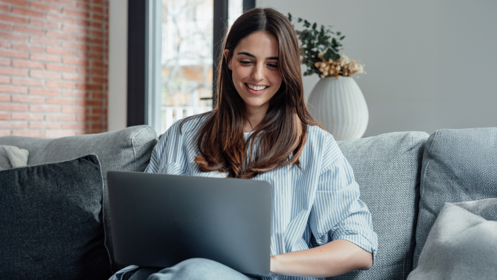 A woman with long brown hair sits on a gray couch, smiling as she searches for Etsy shop ideas on a laptop. A vase with flowers is in the background.