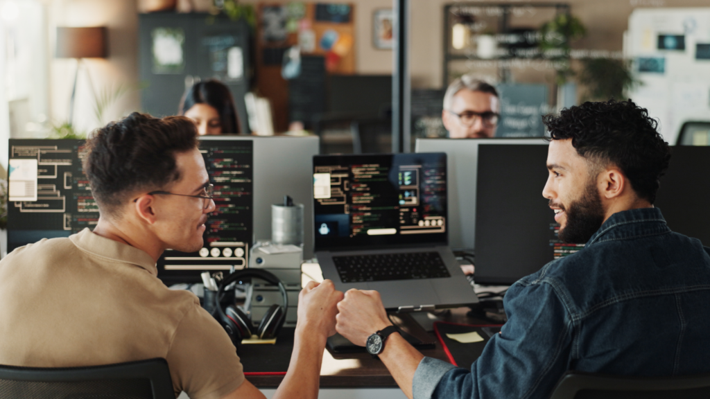 Two men seated at a desk, each working on their own computer, focused on their tasks.