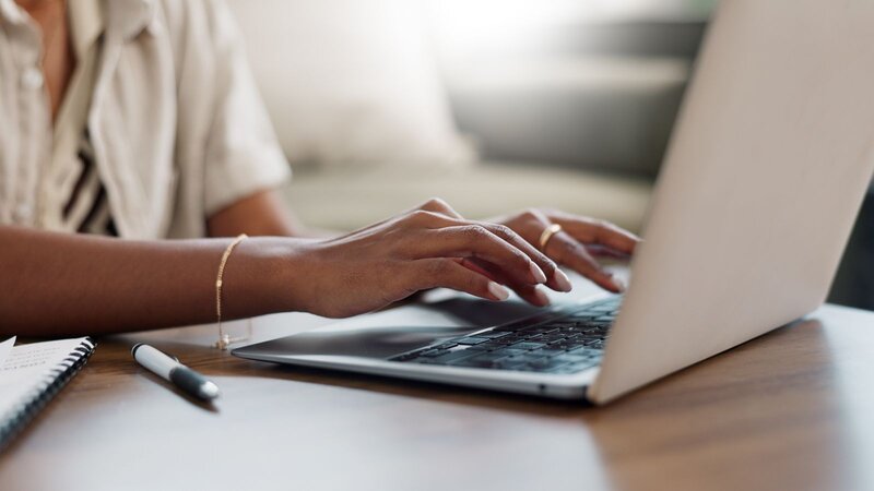 A person typing on a laptop, with a notebook and pen on the table.