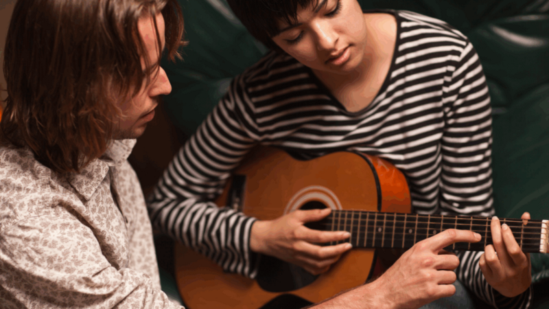 A young man teaching a young woman how to play the guitar.